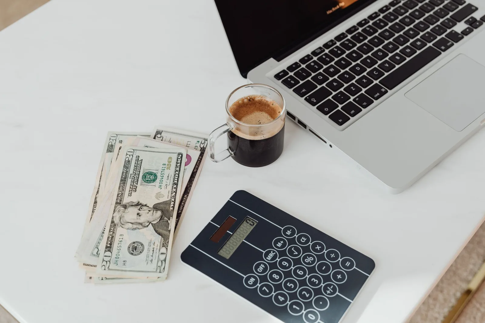 Taschenrechner, Geldscheine und Kaffee neben einem Laptop auf einem weißen Tisch