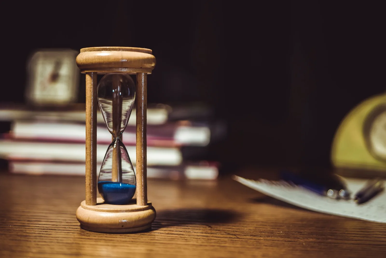 Sanduhr aus Holz mit blauem Sand auf einem Schreibtisch, dahinter Bücher