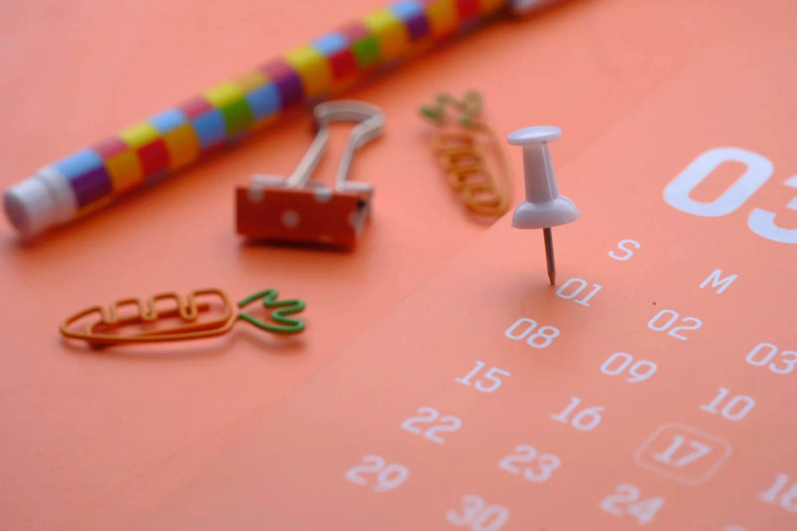 Orangefarbener Kalender mit Pinnnadel, Büroklammern und buntem Stift