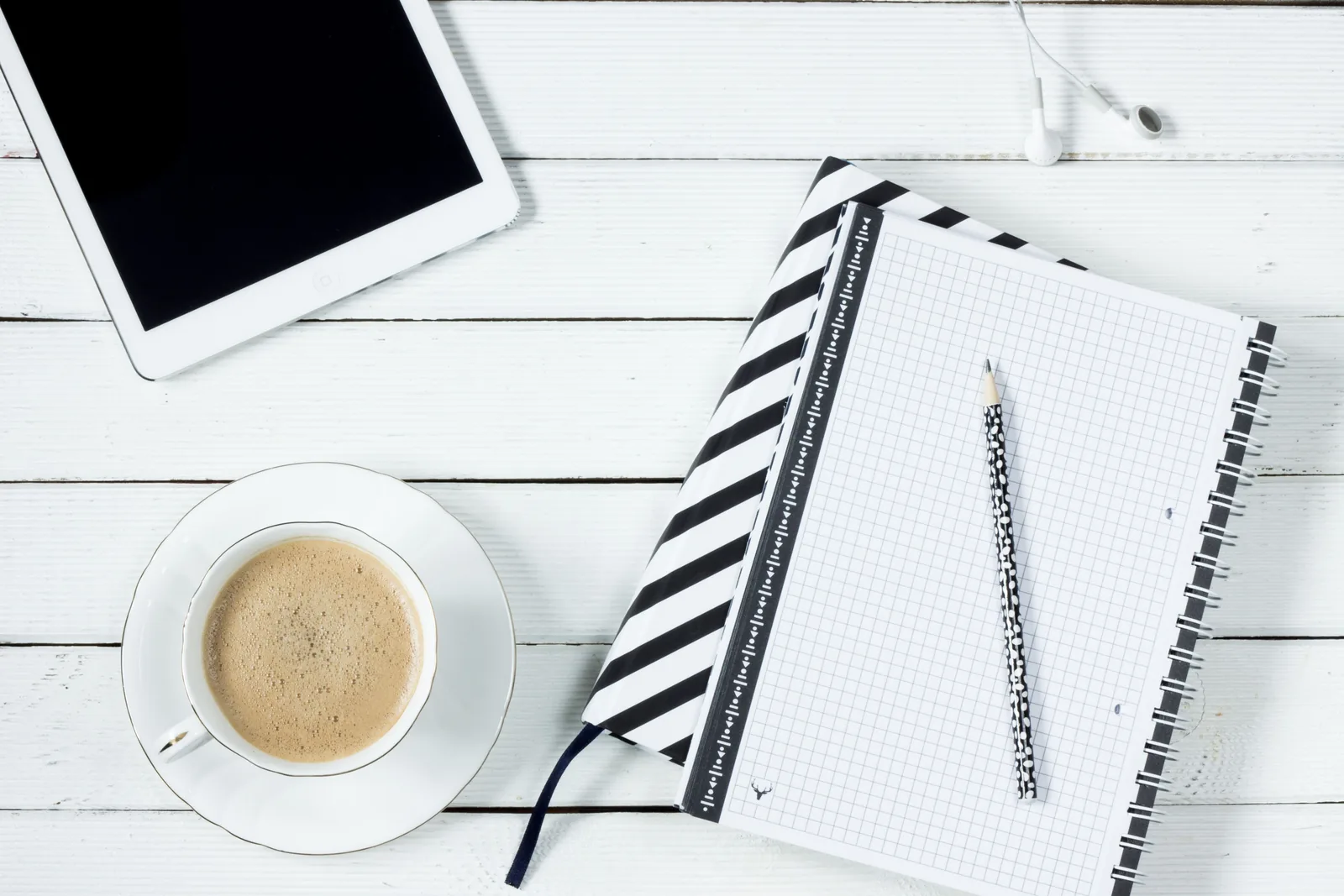 Tablet, Kaffeetasse und kariertes Notizbuch mit Bleistift auf einem weißen Holztisch