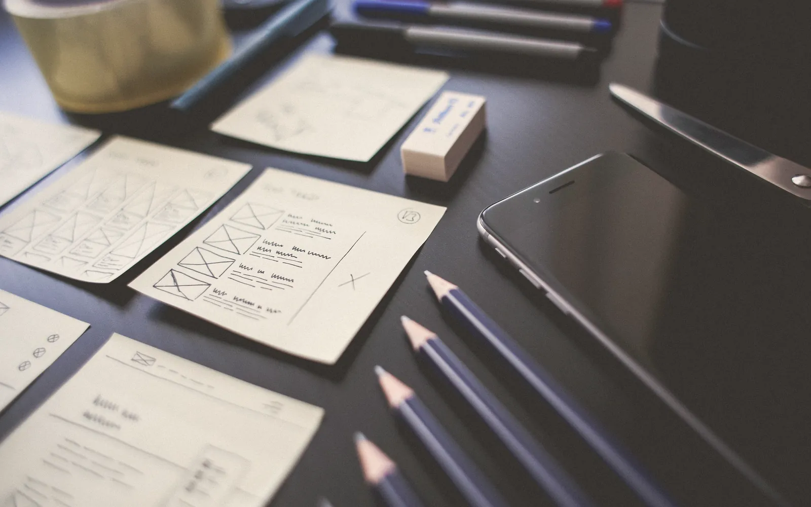 Skizzierte Wireframes, Stifte und Smartphone auf einem dunklen Tisch